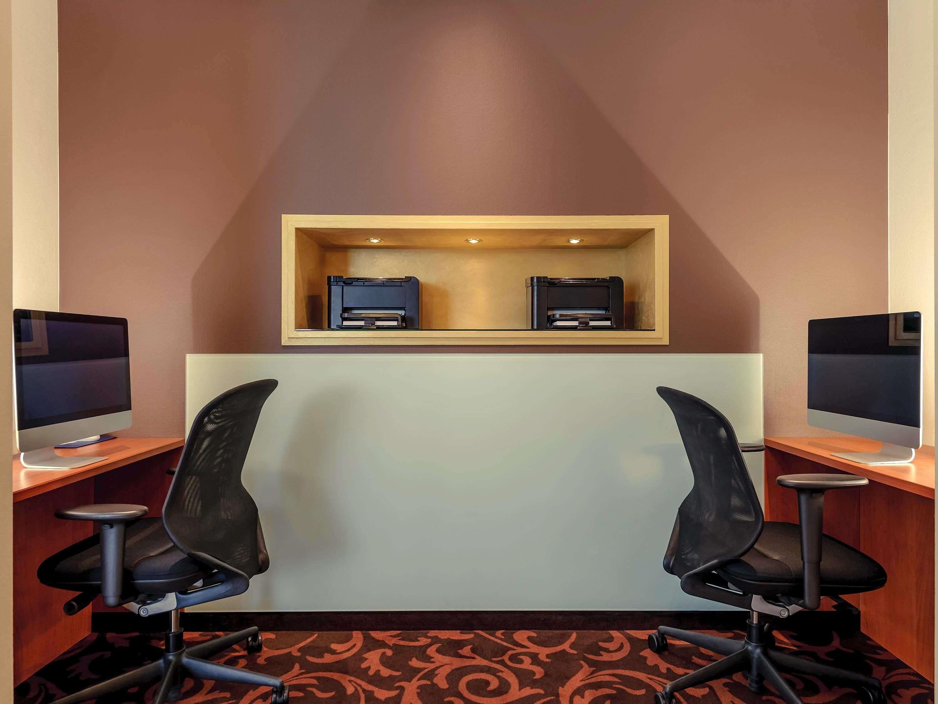 Habitación de hotel con dos escritorios, cada uno con un monitor de computadora e impresora. Hay dos sillas de oficina y piso alfombrado.