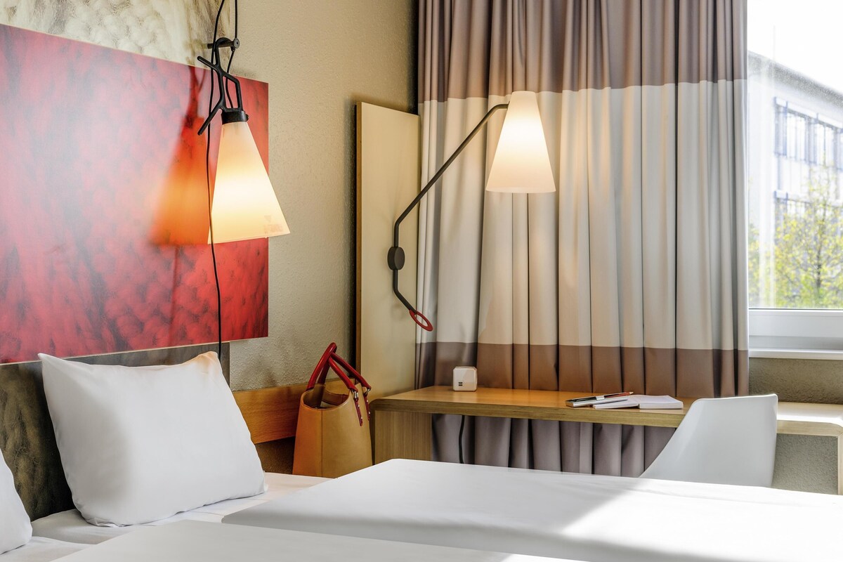 A hotel room with a bed, a desk, a lamp, a handbag, and a window with curtains.