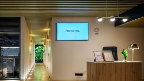 A hotel reception area with a reception desk, a digital screen displaying 'NOVOTEL,' and a wooden reception desk with a lamp and framed certificates. - Novotel Krakow Centrum (Kraków)