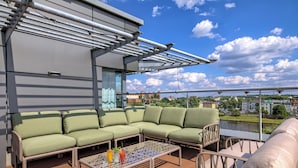 A rooftop terrace with a green sofa, a coffee table with drinks, and a view of a river and buildings. - Novotel Krakow Centrum (Kraków)