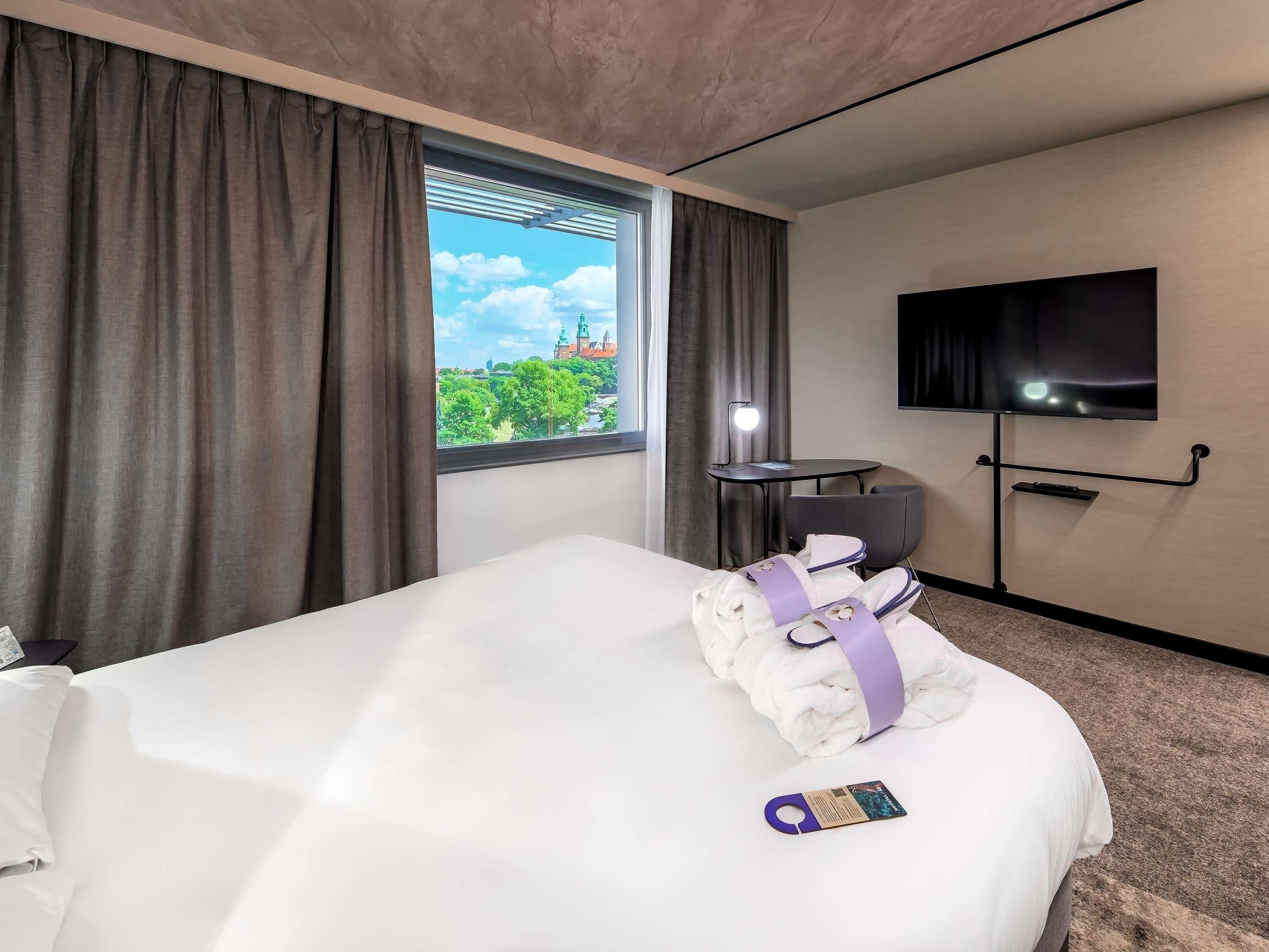 A hotel room with a bed, a TV, a window with curtains, a desk with a lamp, and a view of a cityscape.