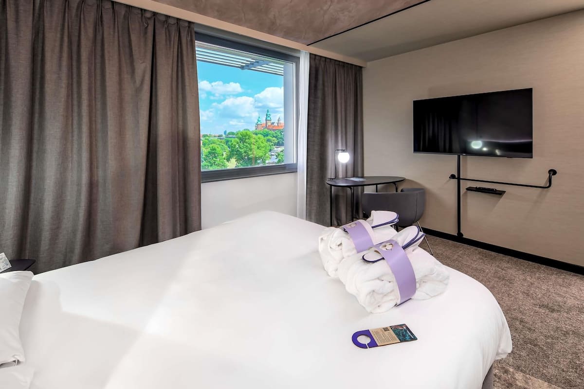 Hotellihuone, jossa on sänky, televisio, ikkuna verhoilla, työpöytä lampun kanssa ja näkymä kaupunkimaisemaan.