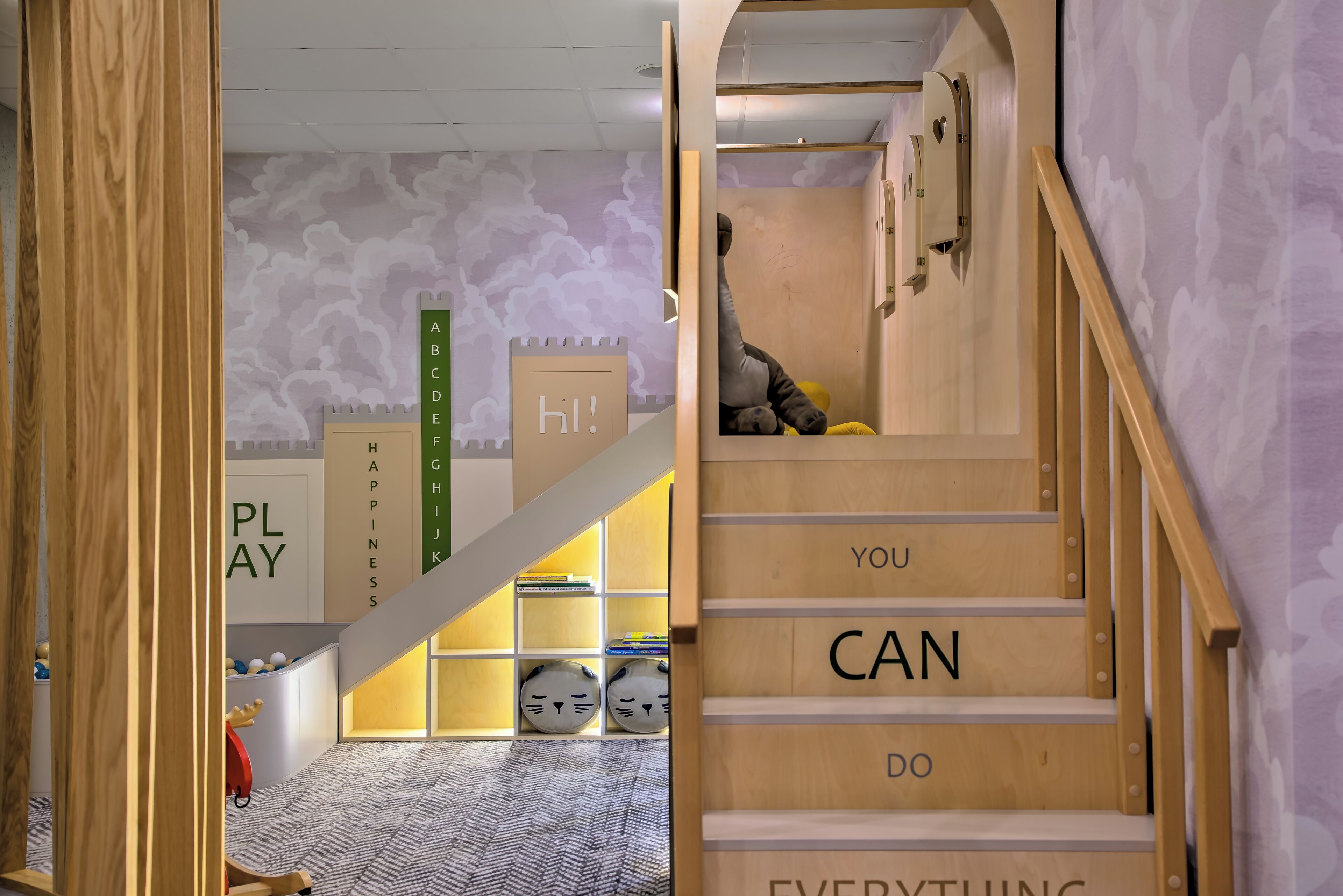 Una escalera de madera con texto motivacional, un área de juegos con letras del alfabeto y un cartel que dice '¡hola!', y una pared con diseño.