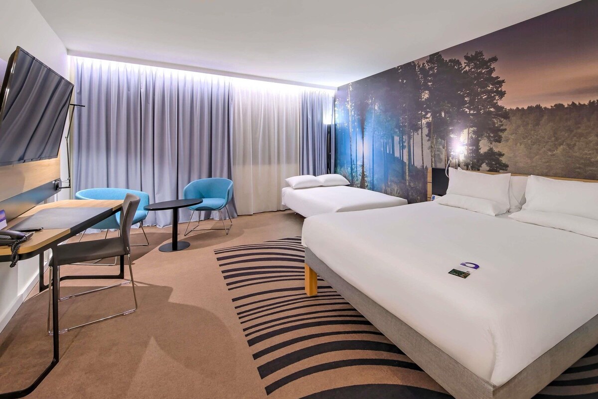A hotel room with a bed, a desk, a chair, and a large window with curtains.