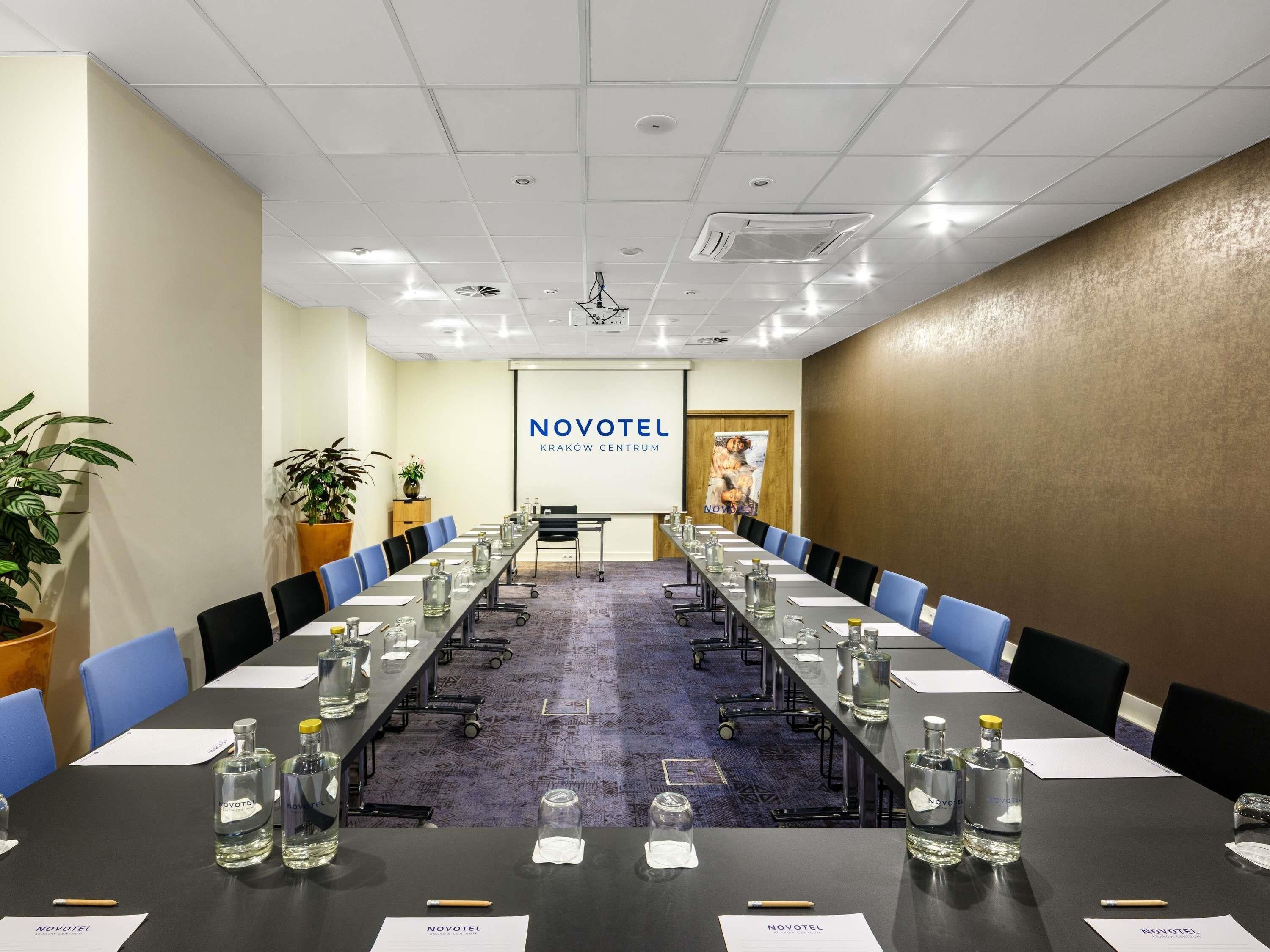Un salón de conferencias con una larga mesa, sillas, botellas de agua y una pantalla de proyector que muestra 'NOVOTEL'.