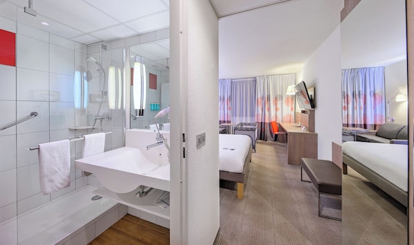 A modern hotel room with a bathroom, a desk, a bed, and a small seating area.