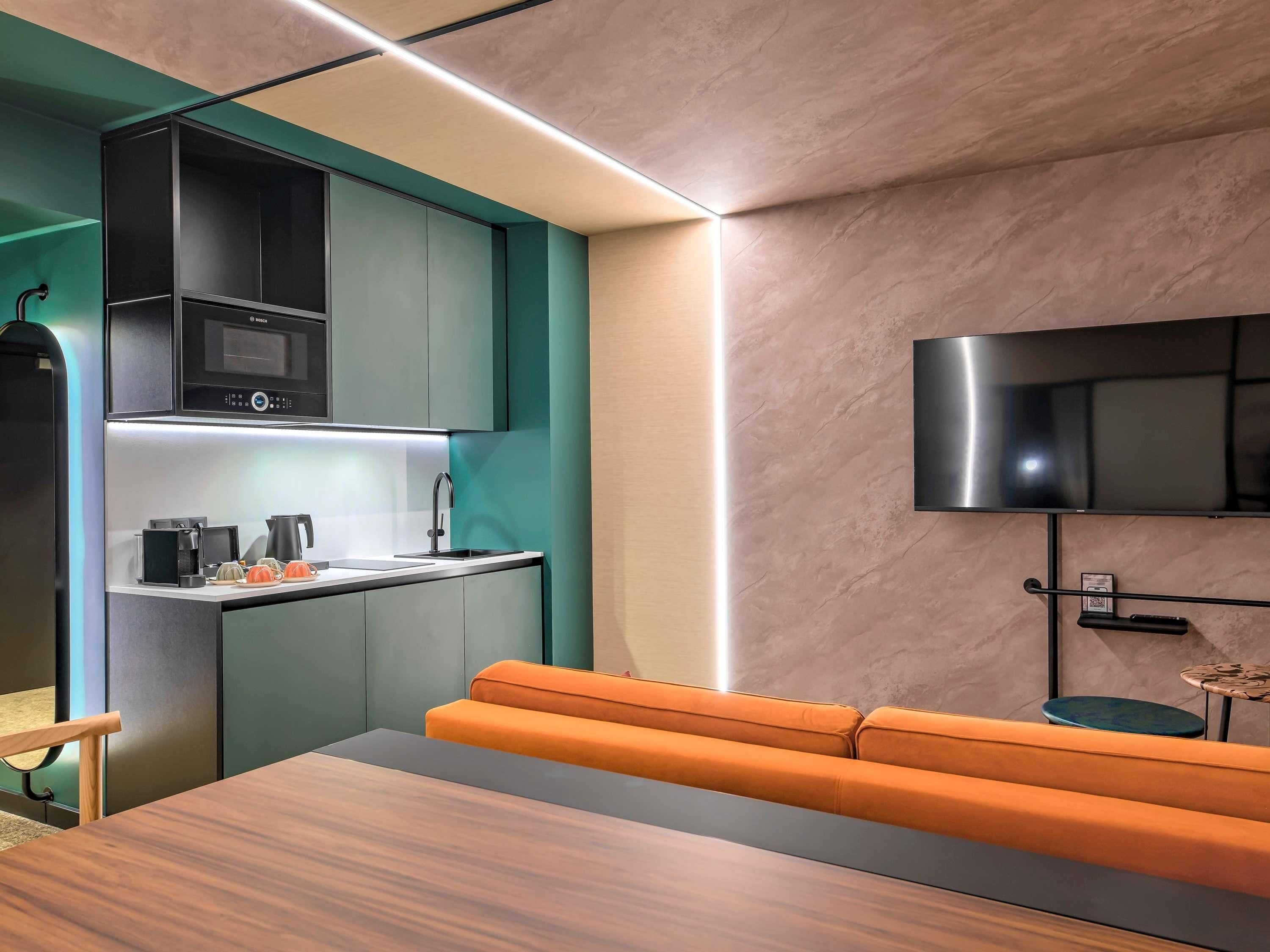 A modern kitchen with built-in appliances, a flat-screen TV mounted on the wall, and a seating area with a wooden table and orange cushions.