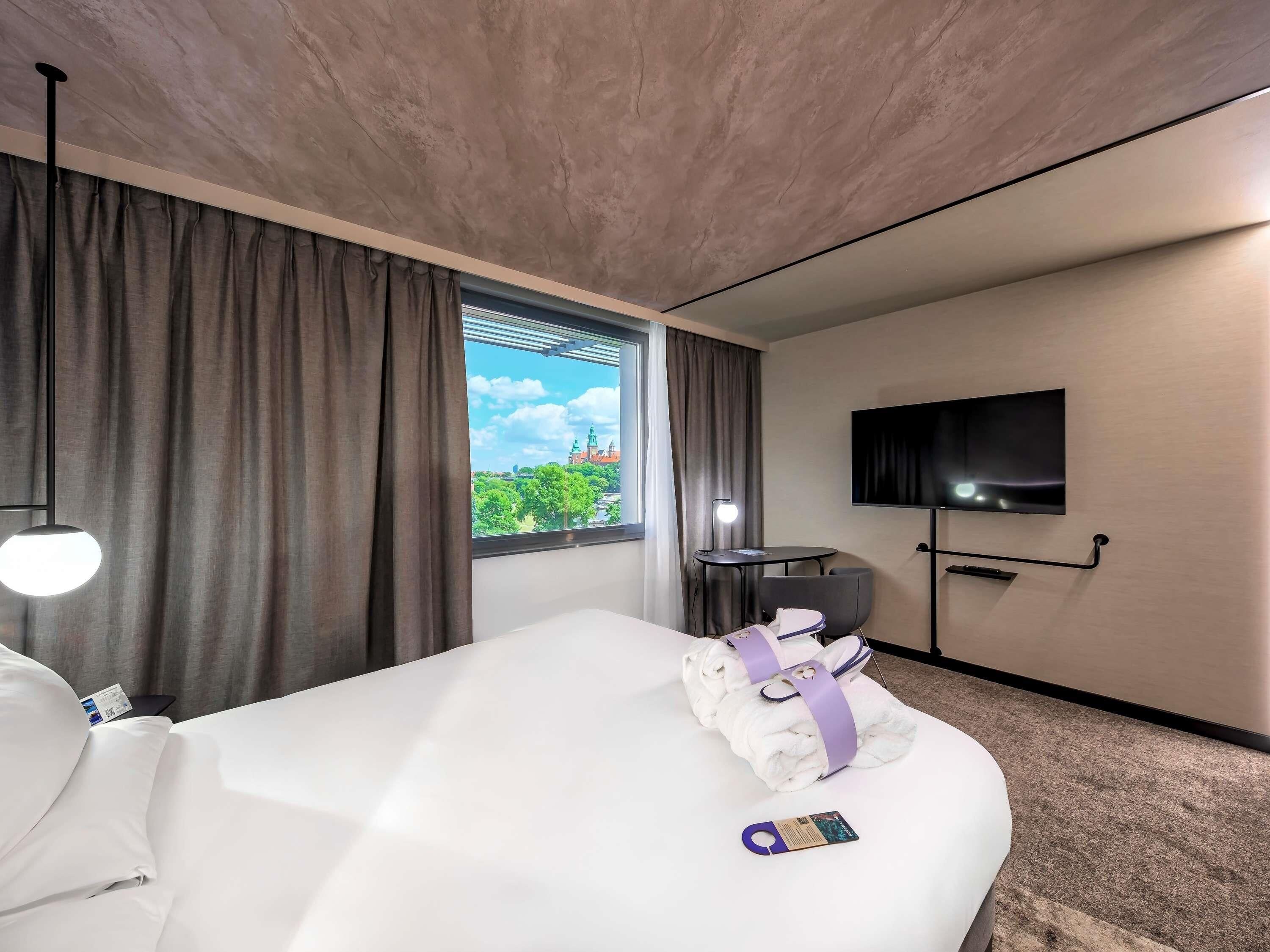 A hotel room with a bed, a TV, a window with curtains, and a view of a building.