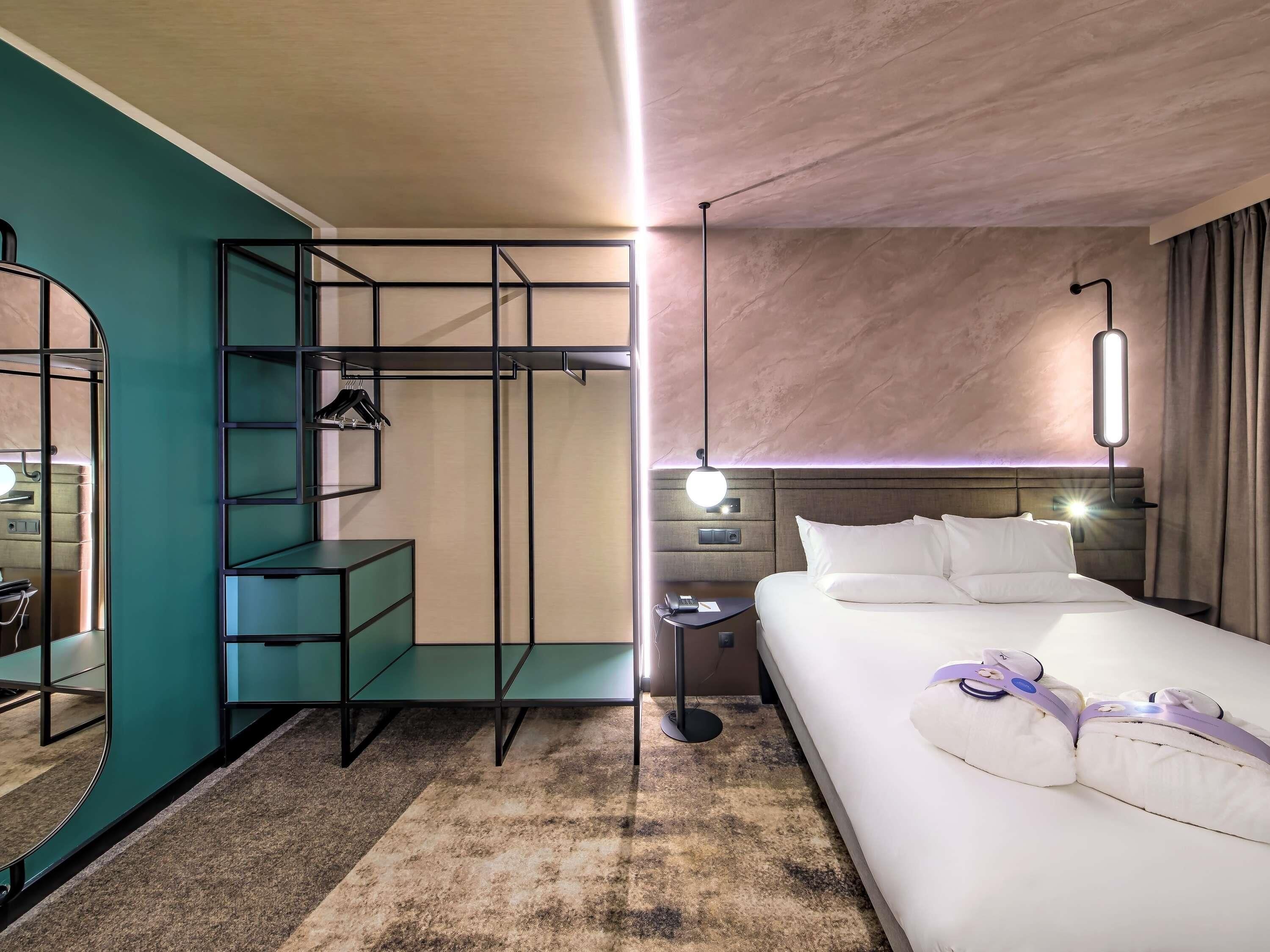 A hotel room with a bed, a desk with a chair, a wardrobe, a mirror, a nightstand, and a bedside table.