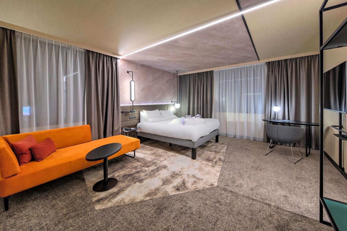 A modern hotel room with a bed, a sofa, a desk, and a TV.
