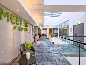 A modern hotel lobby with a green wall displaying 'MEETING AT NOVOMOTEL,' a seating area with a plant, and a patterned carpet. - Novotel Krakow Centrum (Kraków)