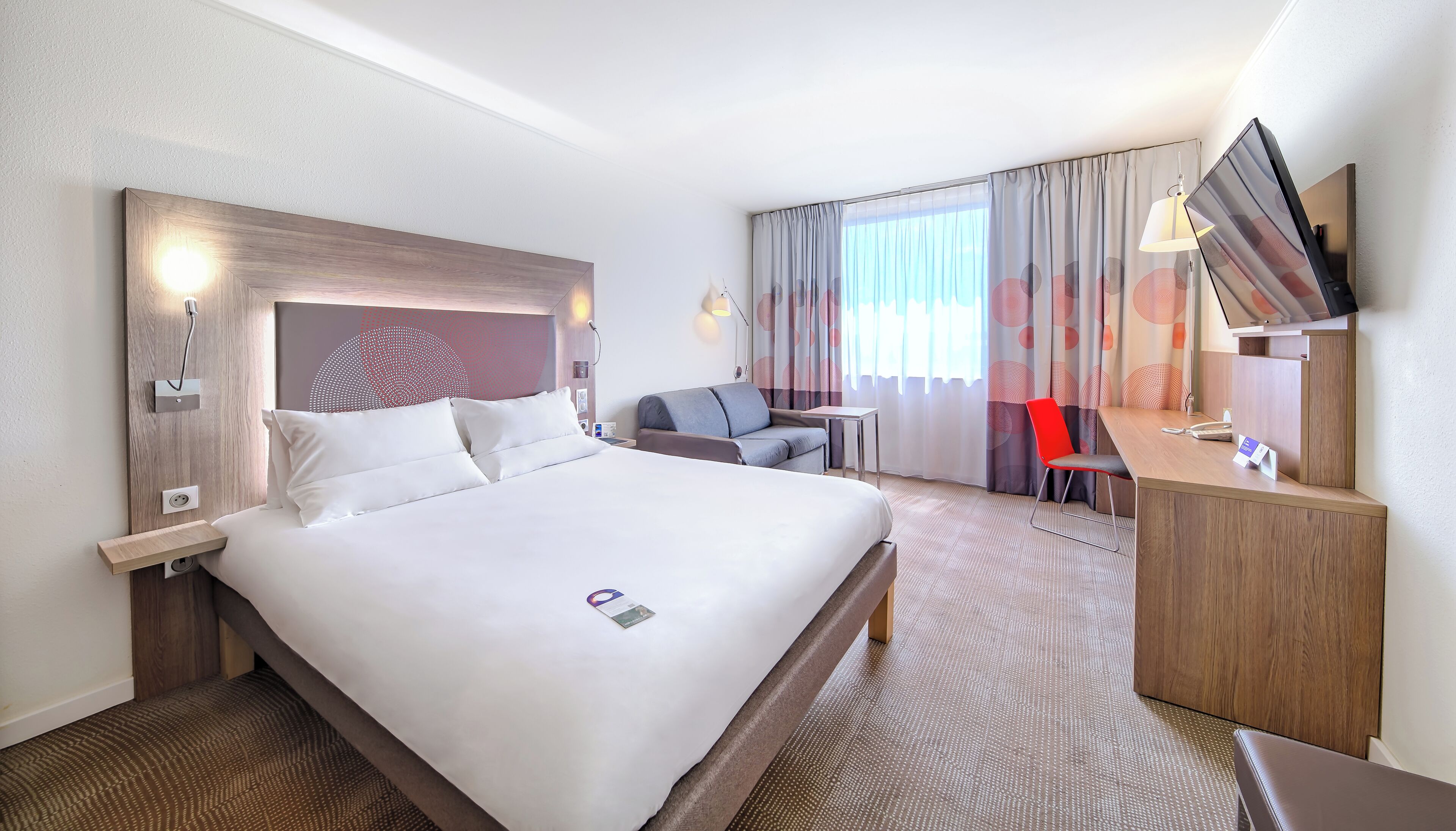 A hotel room with a large bed, a desk, a chair, a TV, and a window with curtains.