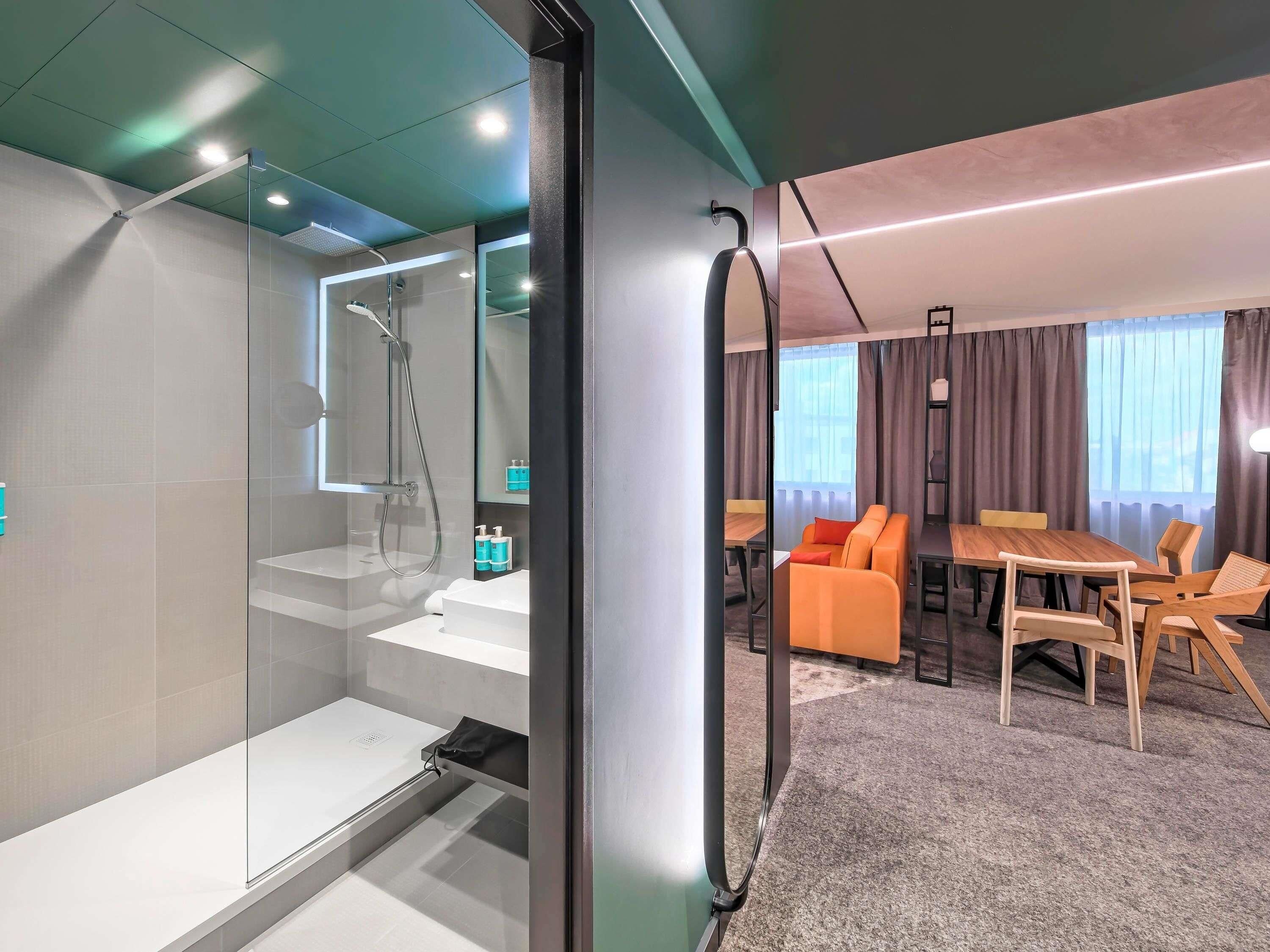 A modern hotel room with a glass shower enclosure, a wooden dining table, and a sofa.