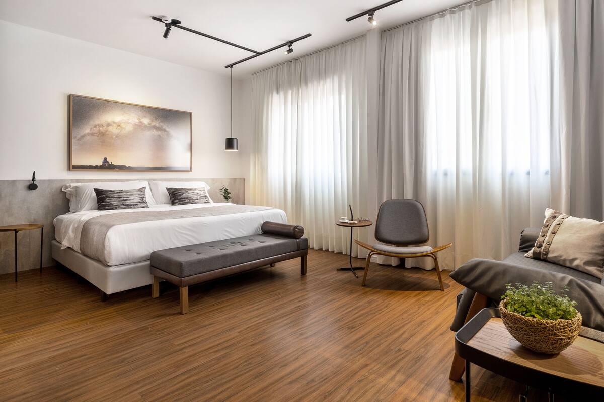 Une chambre d’hôtel moderne dotée d’un grand lit, d’un coin salon avec un fauteuil et une petite table, d’un canapé et d’une grande fenêtre avec des rideaux.