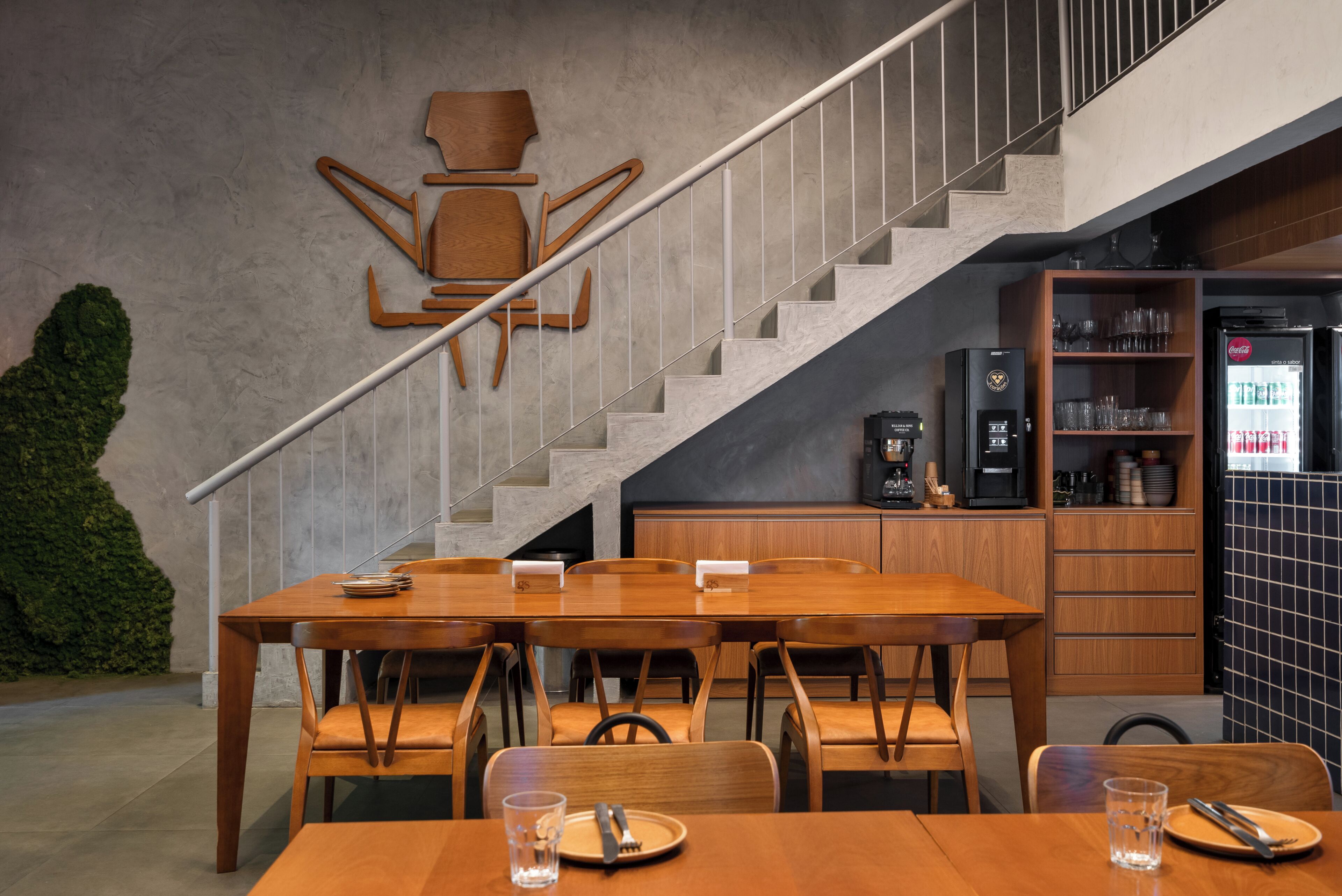 A modern dining area with a wooden table and chairs, a staircase, and a kitchen area with a refrigerator and cabinets.