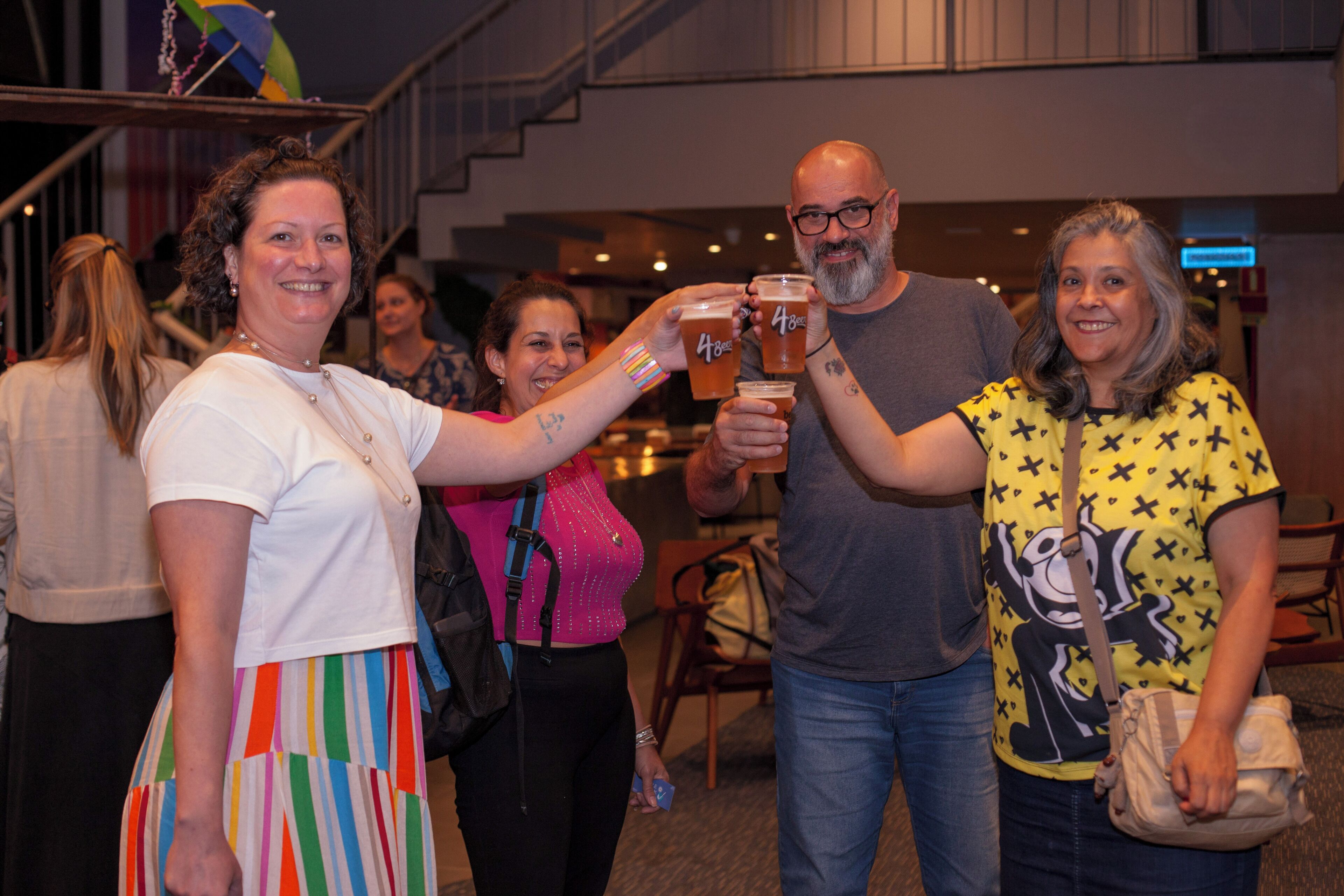 Un grupo de personas brindando con vasos de cerveza en un espacio interior bien iluminado, con escaleras visibles y un letrero al fondo.