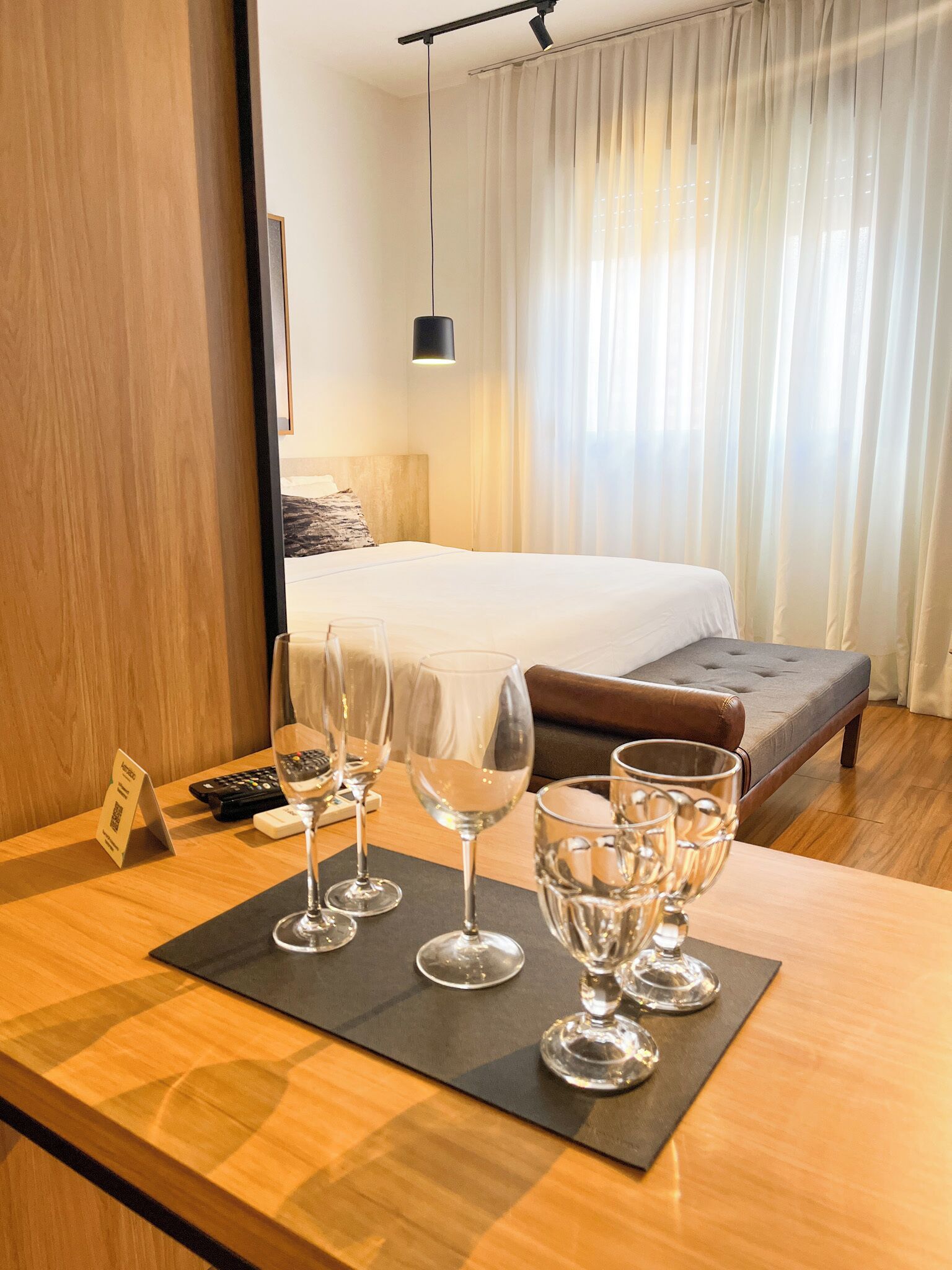 A hotel room with a bed, a bedside table with wine glasses, a remote control, and a sign.
