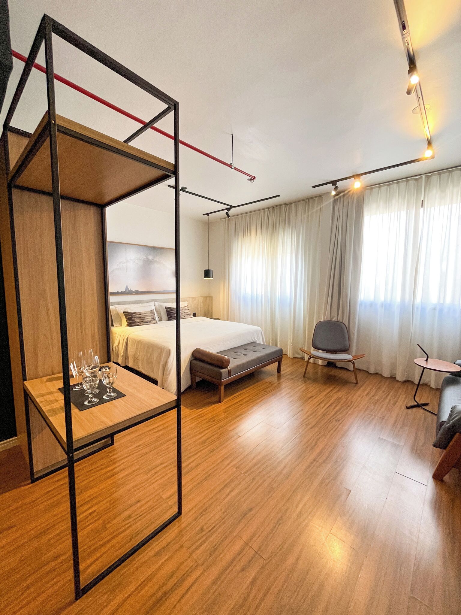 Quarto de hotel com cama, mesa de cabeceira com copos, uma cadeira, uma pequena mesa e uma janela grande com cortinas.
