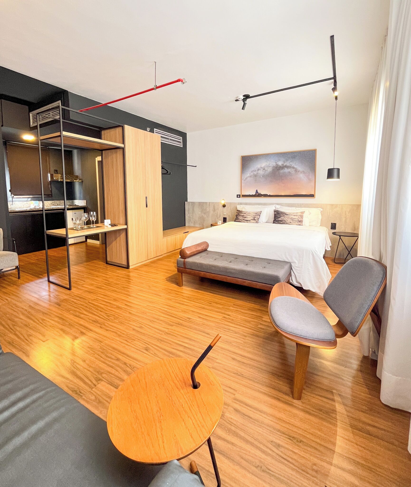 A modern hotel room with a bed, a seating area with a table, a wardrobe, and a kitchenette.