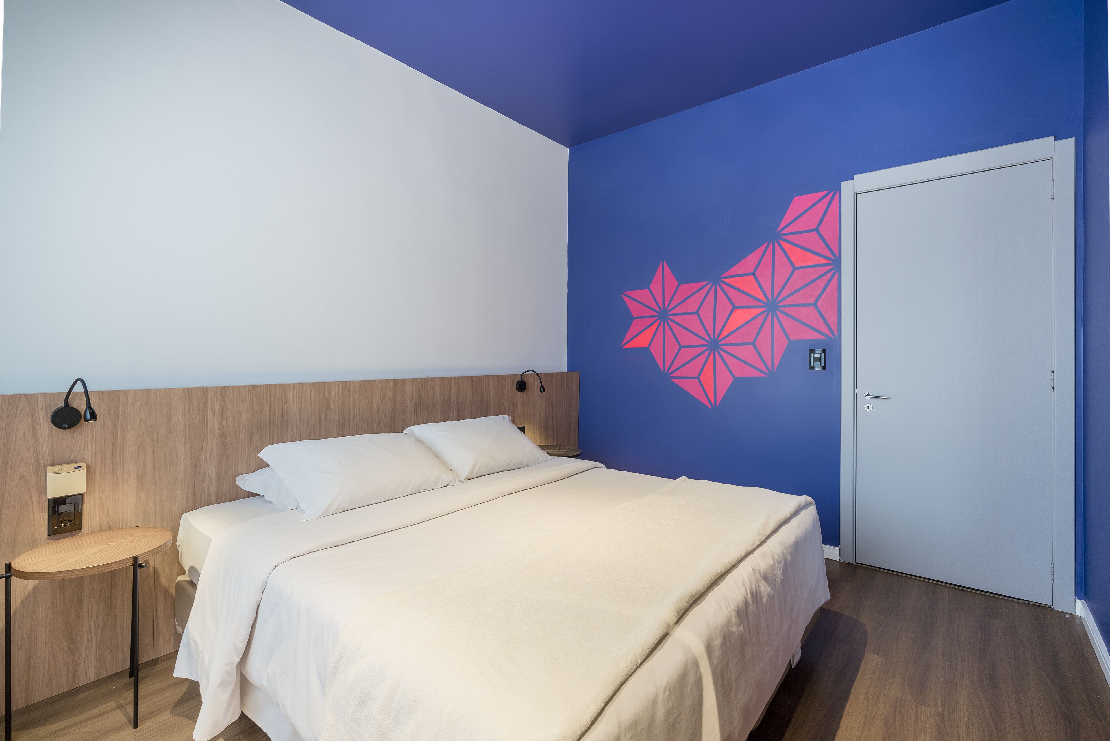 A bedroom with a bed, a bedside table, a wall-mounted lamp, and a door.