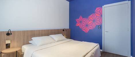 A bedroom with a bed, a bedside table, a wall-mounted lamp, and a door.