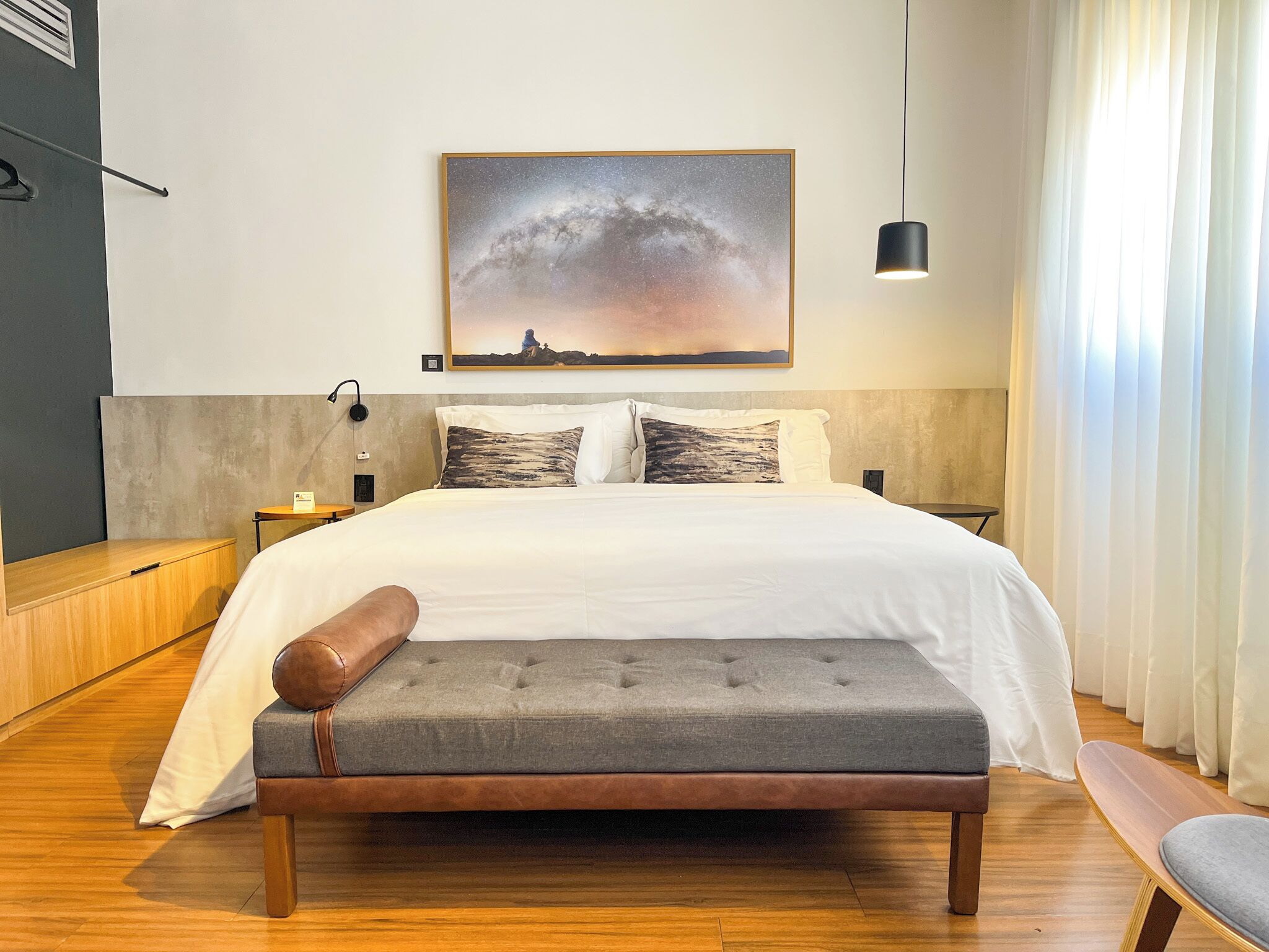 Una habitación de hotel moderna con una cama grande, un sofá gris, suelo de madera y un cuadro de un paisaje en la pared.