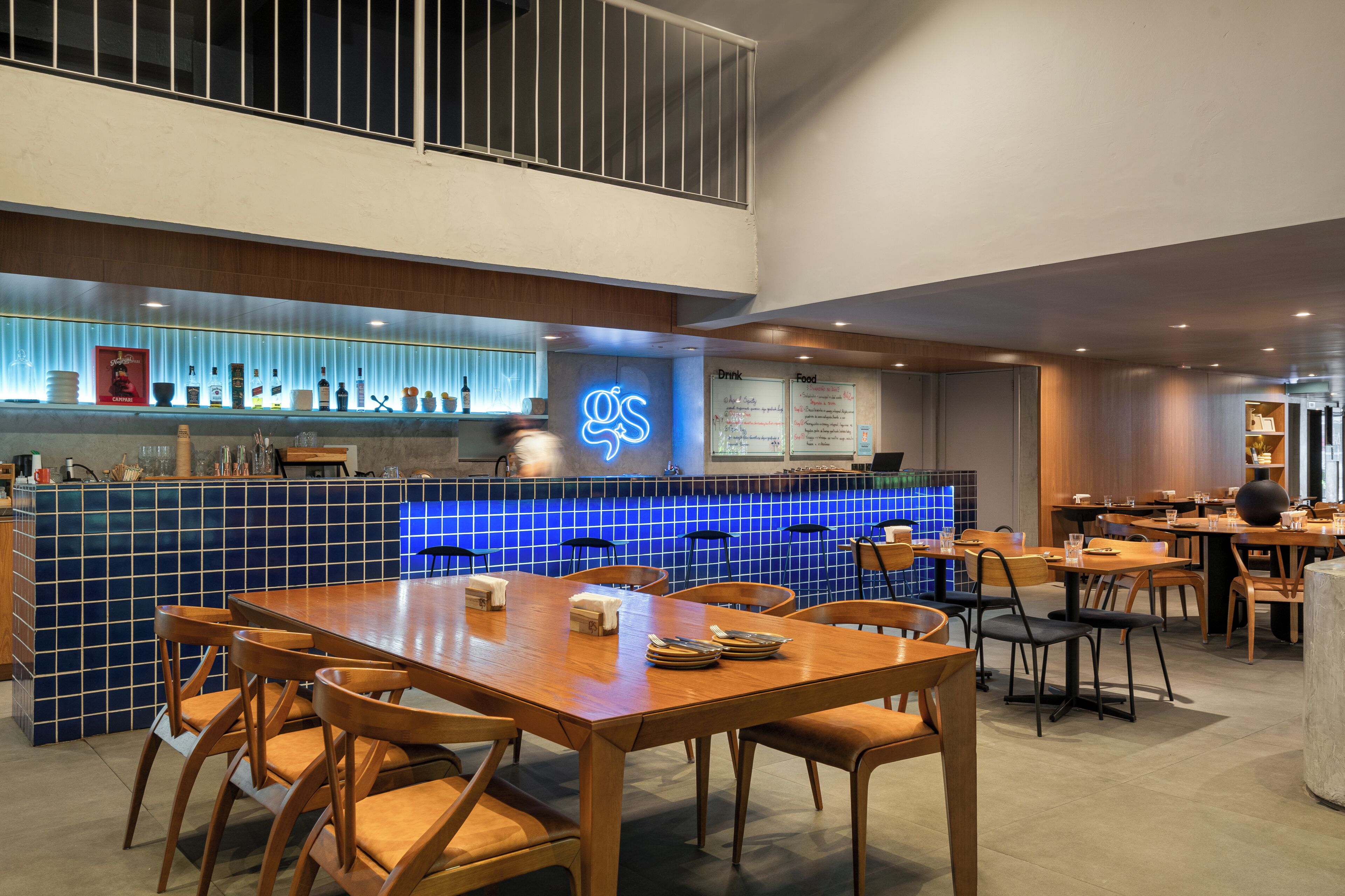 En modern restaurang med en bardisk klädd i blått kakel, bord och stolar i trä samt en neonskylt.