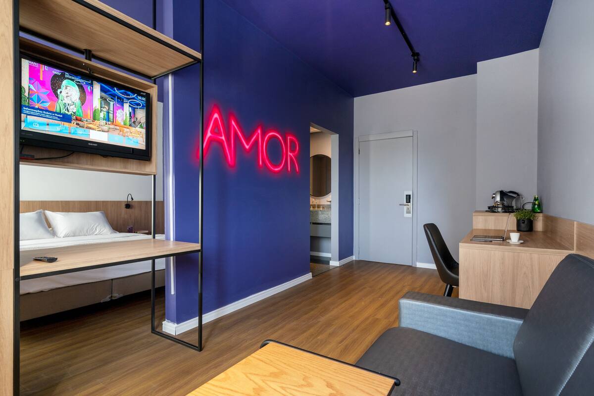 Une chambre d’hôtel moderne équipée d’une télévision, d’un néon lumineux affichant le mot « AMOR », d’un bureau et d’un lit.