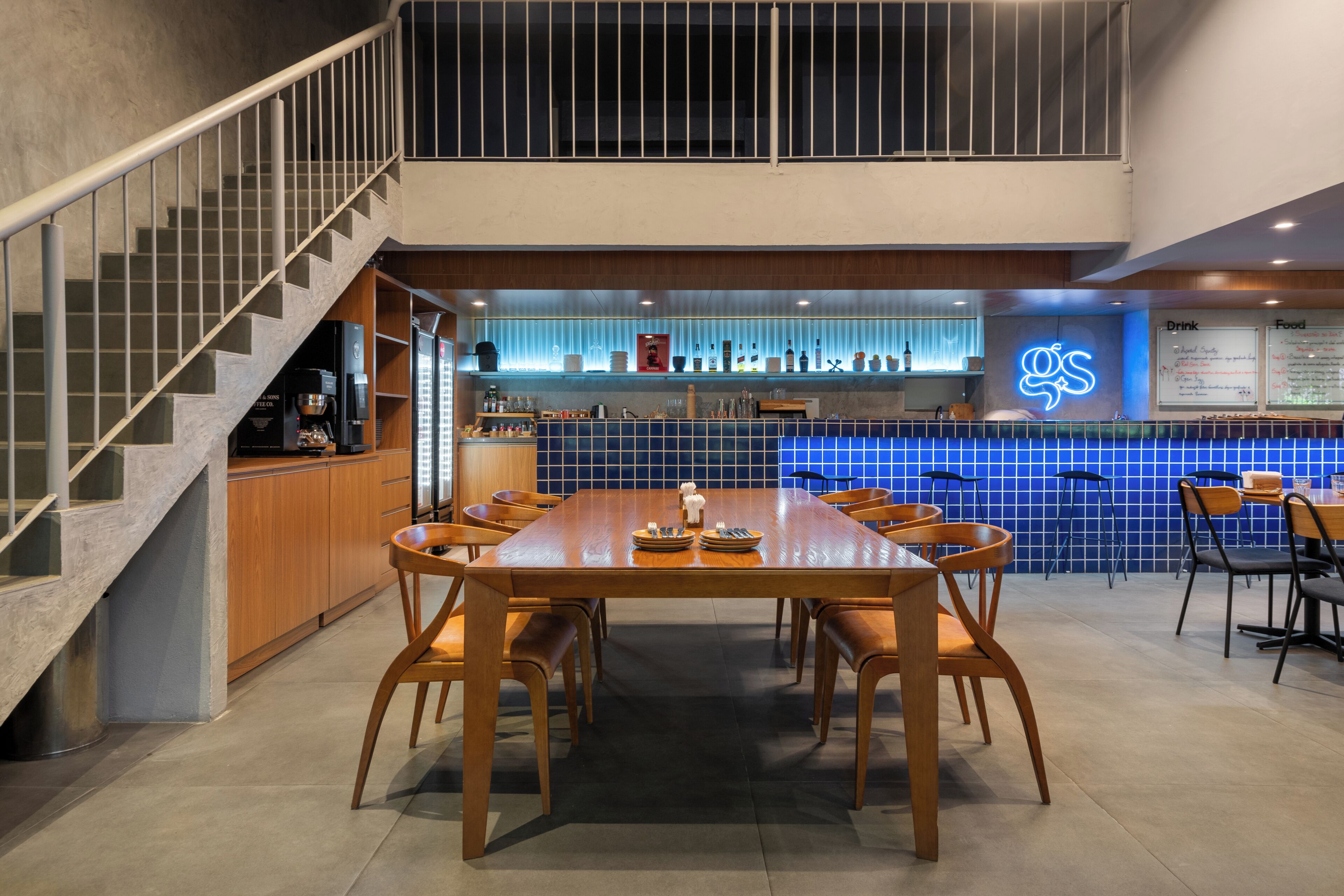 A modern interior with a wooden dining table, chairs, a staircase, and a bar area with a neon sign.