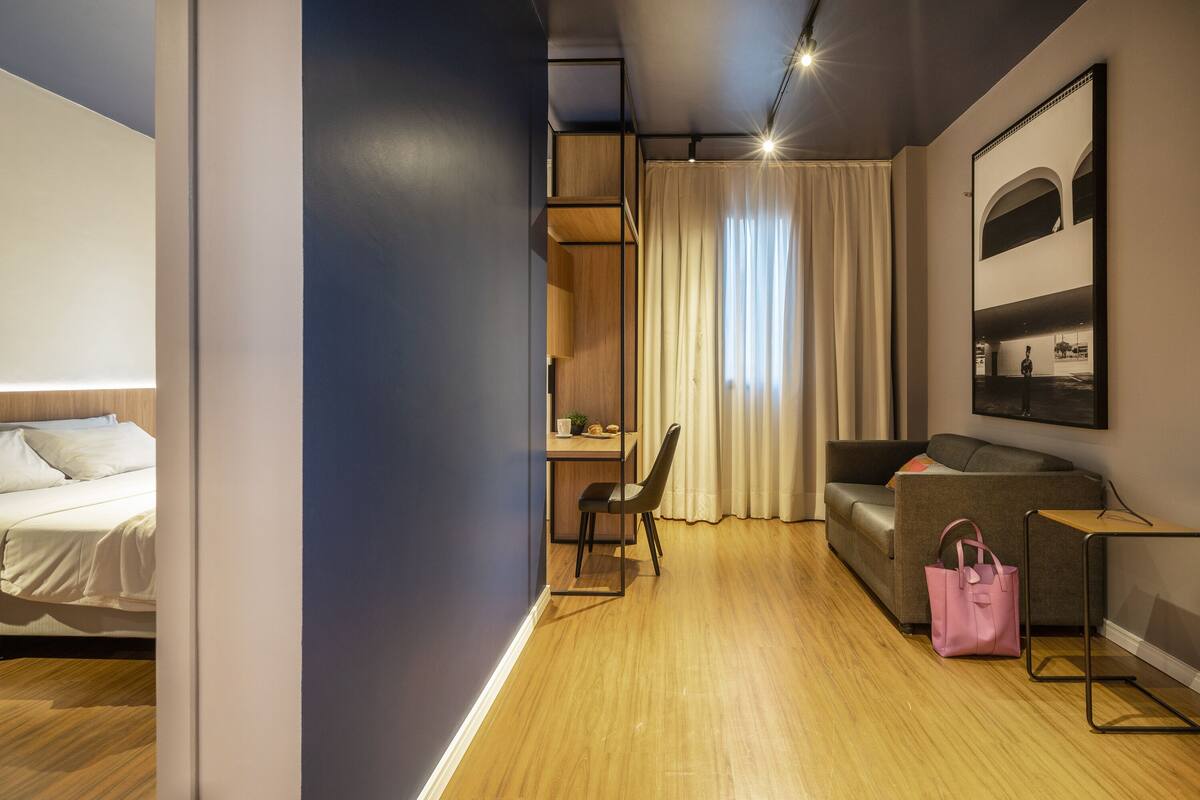 Une chambre d’hôtel avec un lit, un bureau, une chaise, un canapé, un sac rose et un tableau encadré au mur.