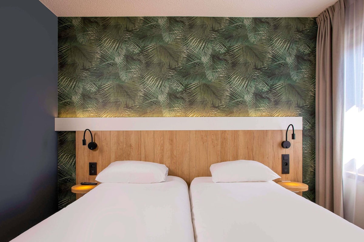 Quarto de hotel com cabeceira de madeira, duas camas, mesas de cabeceira e luminárias fixadas na parede.