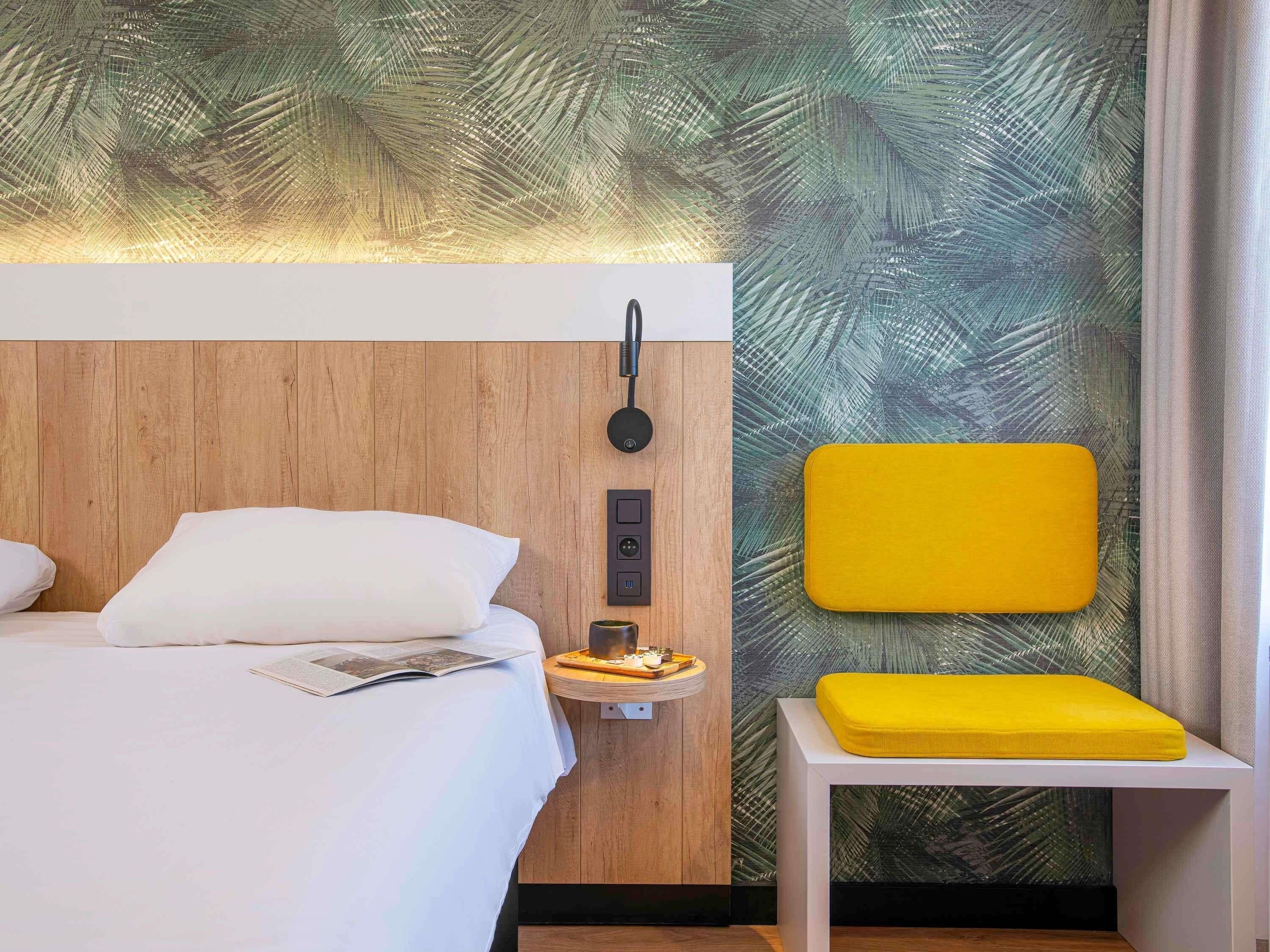 A hotel room with a wooden headboard, a bedside table with a lamp, a yellow chair, and a wall with a leaf pattern.