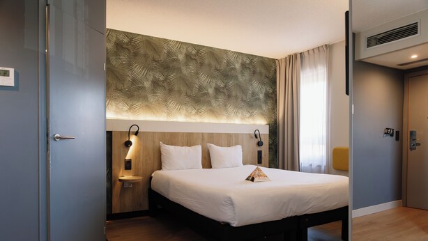 A hotel room with a bed, bedside lamps, a nightstand, a wall-mounted light, and a window with curtains. - ibis Brussels Waterloo (Waterloo)