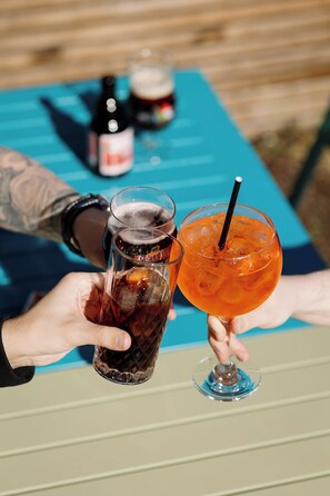 Two people holding drinks, one in a glass and one in a bottle, with a table and a bottle in the background.