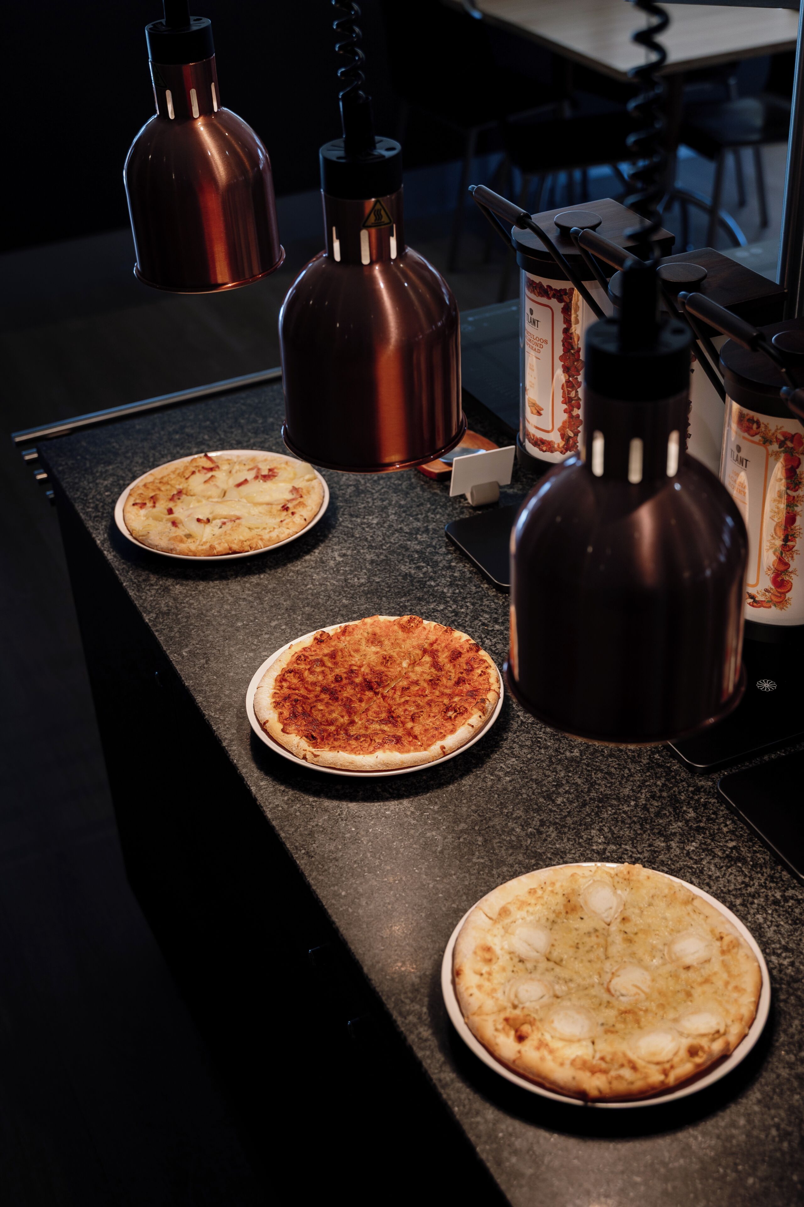 A kitchen counter with three pizzas and two copper sauce dispensers.