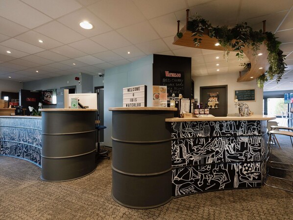 A reception area with a counter, two large barrels, a sign reading 'WELCOME @ IBIS WATERLOO,' and a menu board.