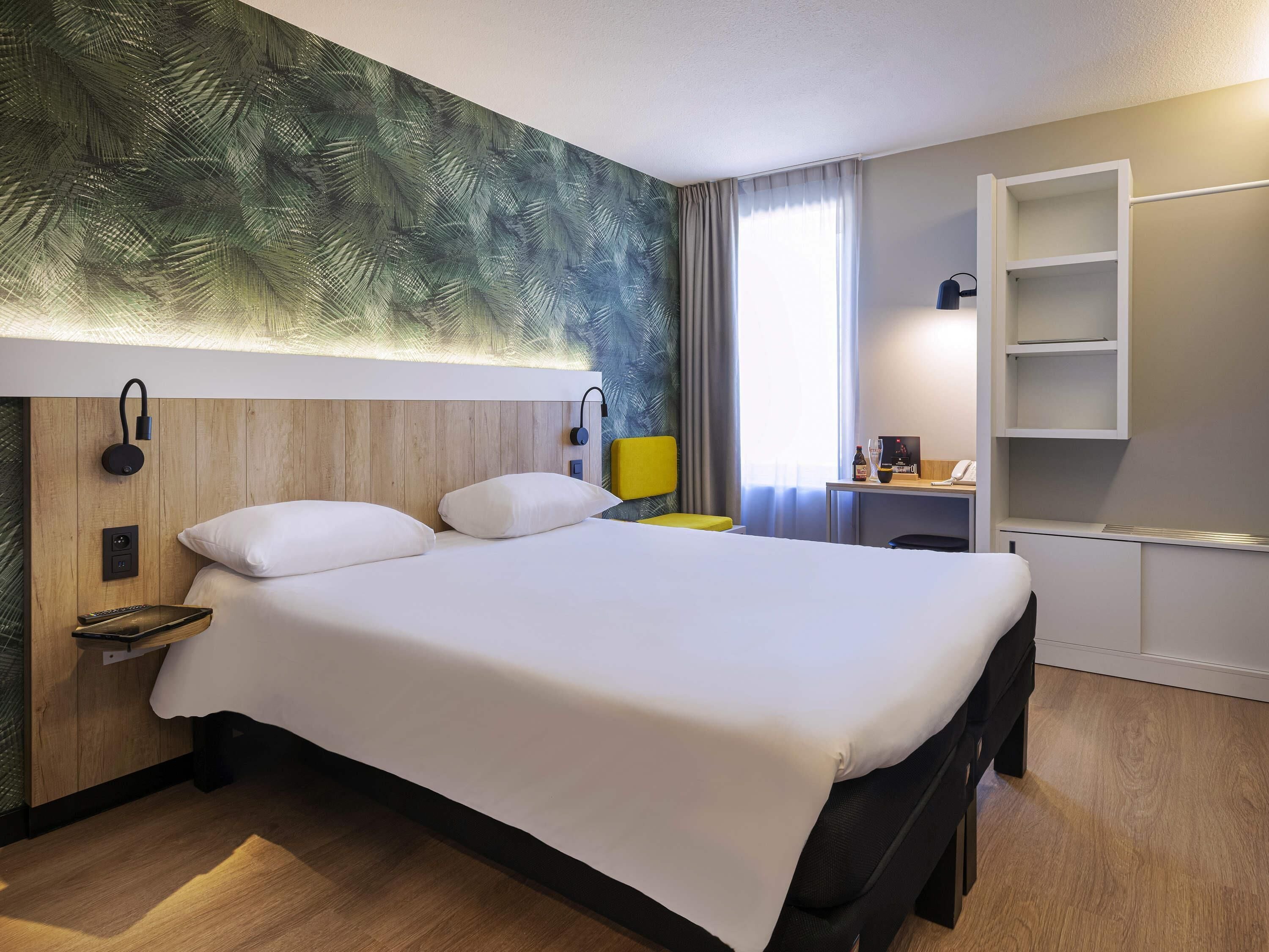 A hotel room with a large bed, a wooden headboard, a desk with a chair, a small table with a lamp, and a built-in shelf.