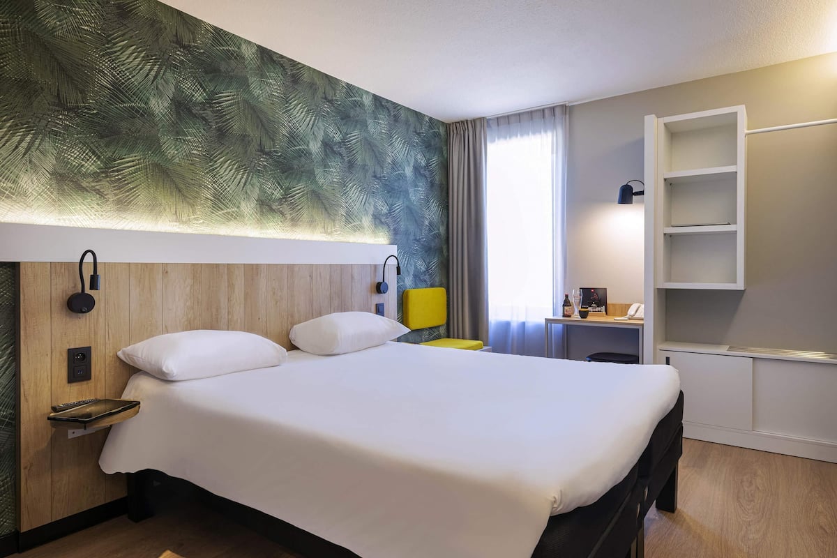 Quarto de hotel com uma cama grande, cabeceira de madeira, escrivaninha com cadeira, pequena mesa com abajur e prateleira embutida.