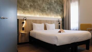 Une chambre d’hôtel avec un lit, des lampes de chevet, une tête de lit en bois, une table de chevet et une fenêtre avec des rideaux.
