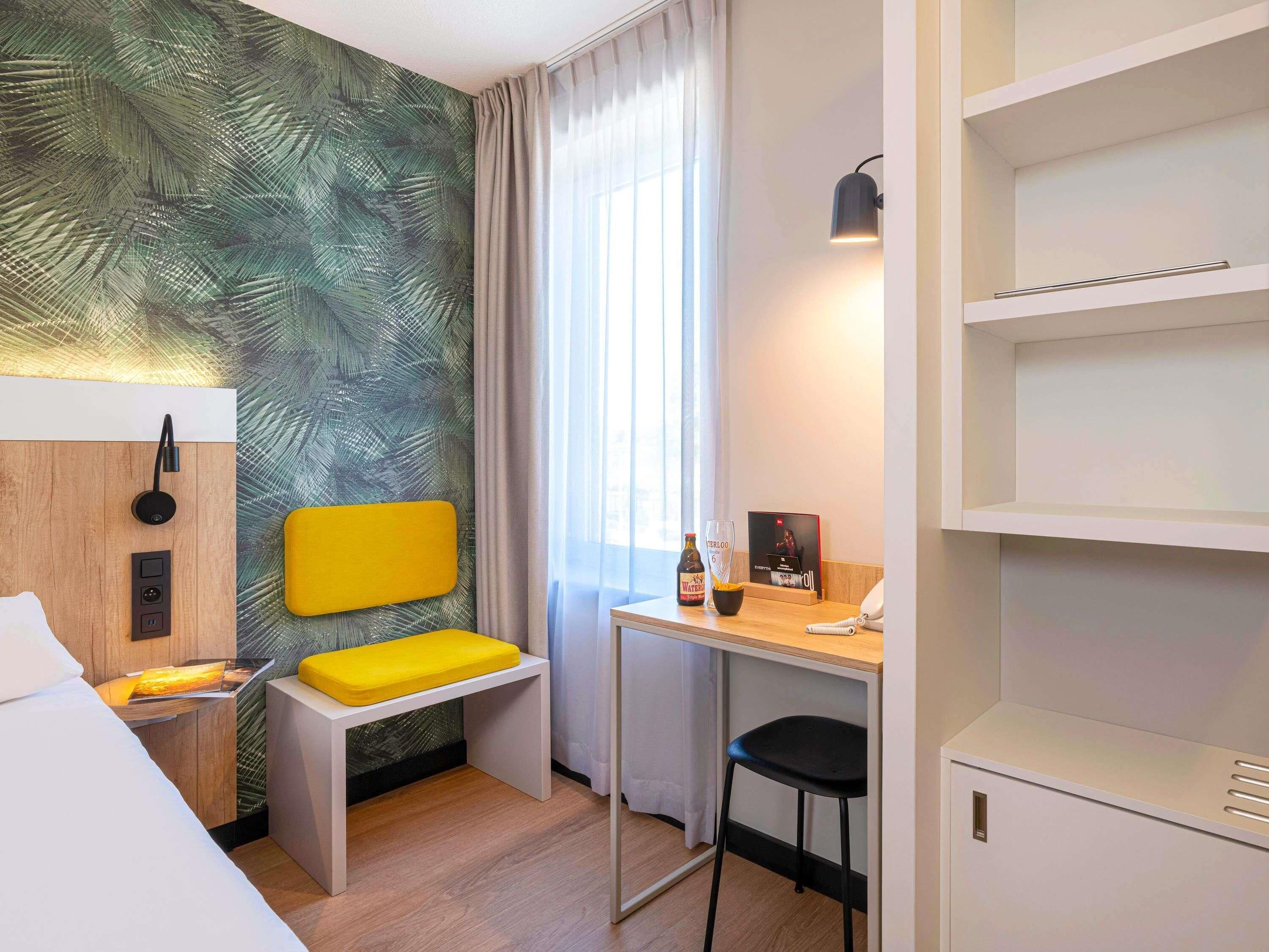 Una habitación de hotel con una cama, un escritorio con silla, una mesita, una lámpara de pared, un teléfono y un estante.