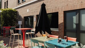 An outdoor patio with tables and chairs, a brick building in the background, and a garden area with flowers. - ibis Brussels Waterloo (Waterloo)
