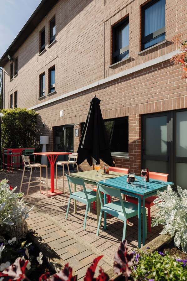 Eine Terrasse im Freien mit Tischen und Stühlen, im Hintergrund ein Backsteinhaus und ein Gartenbereich mit Blumen.