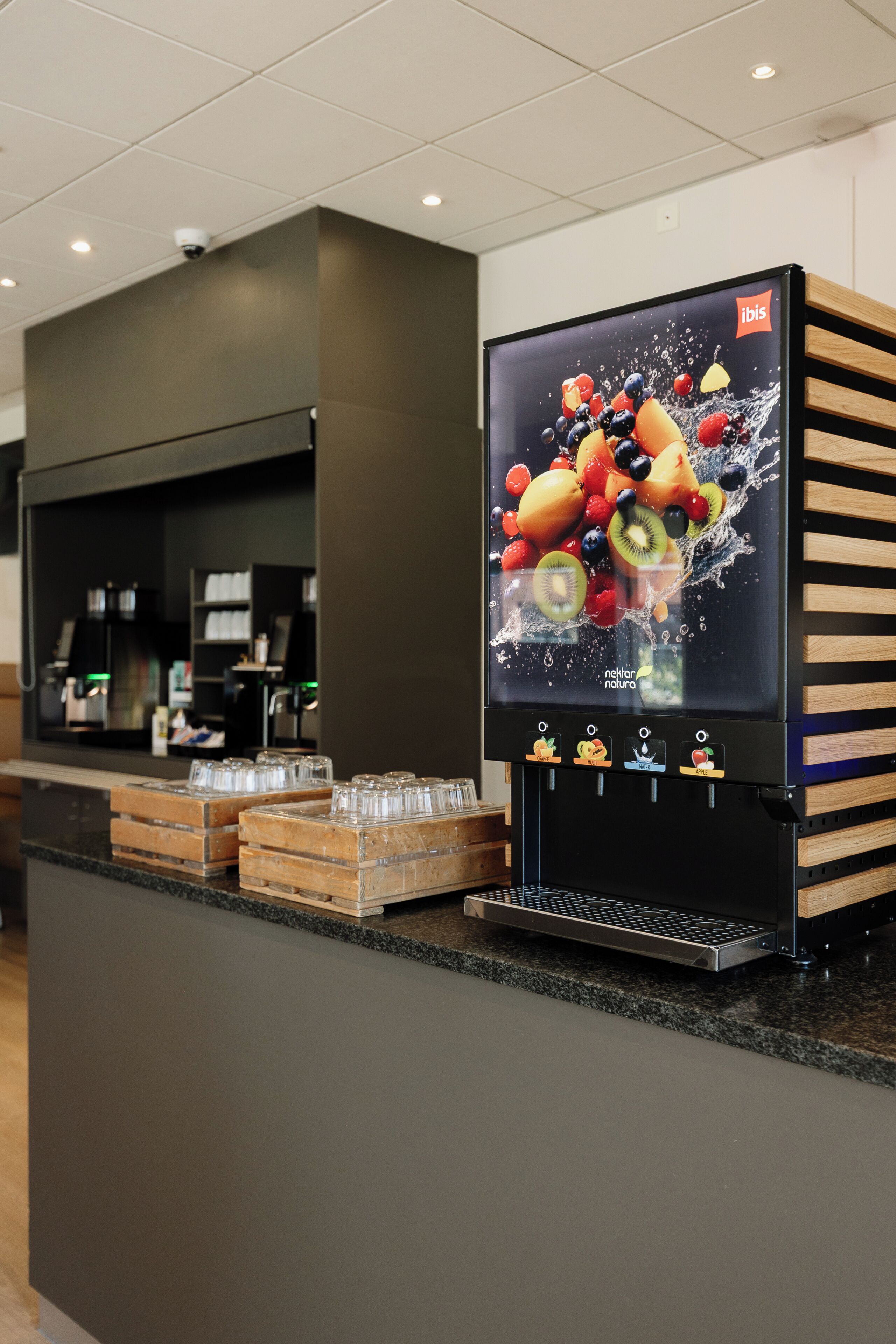Un dispensador de bebidas automático con un gráfico de frutas, una cafetera y cristalería sobre una encimera.
