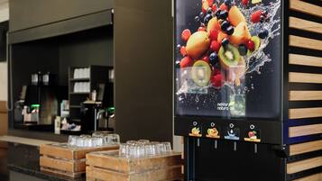 Un dispensador de bebidas automático con un gráfico de frutas, una cafetera y cristalería sobre una encimera.