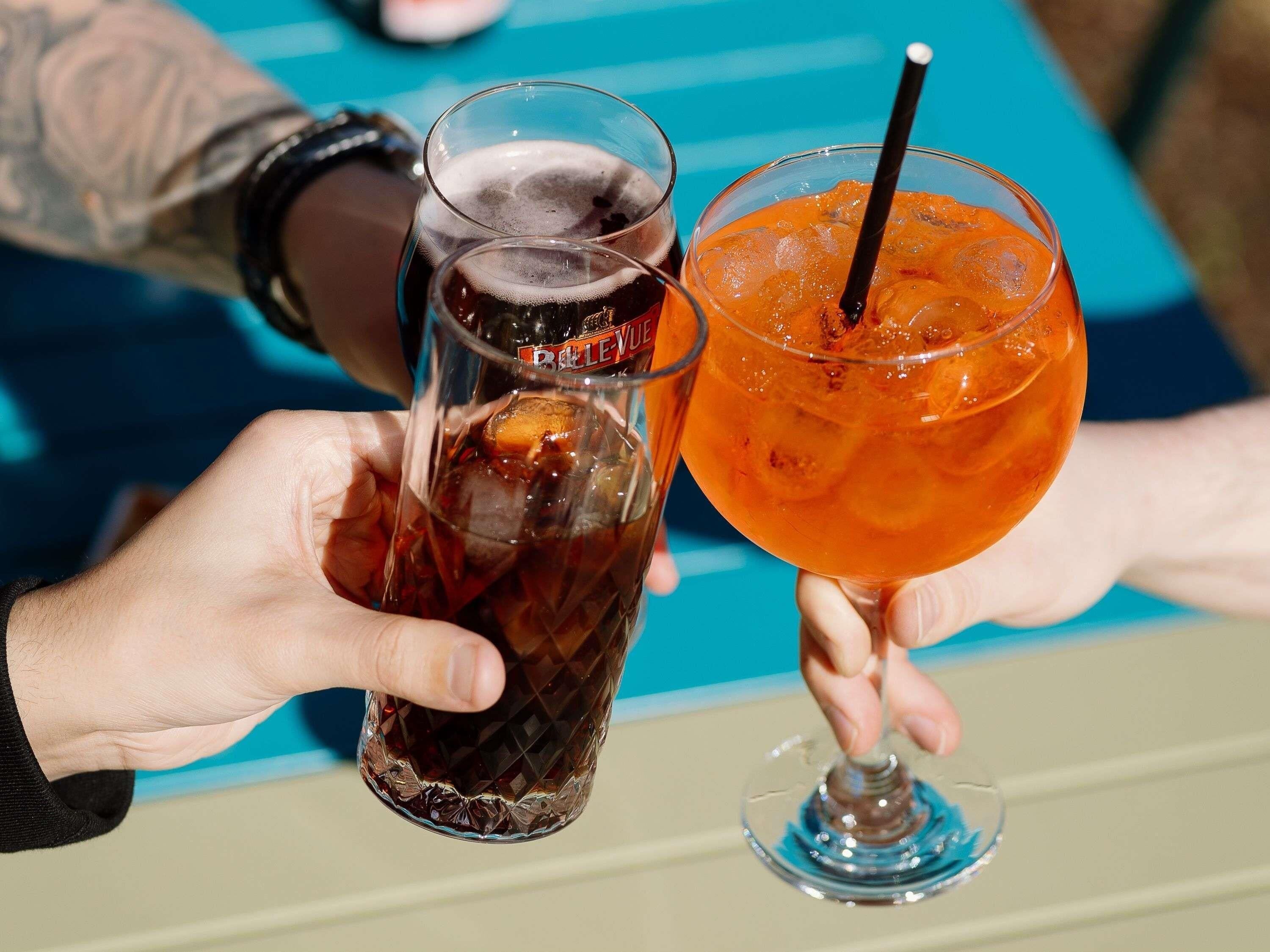 Two hands holding glasses of beverages, one dark and one orange, with a blue-striped tablecloth.