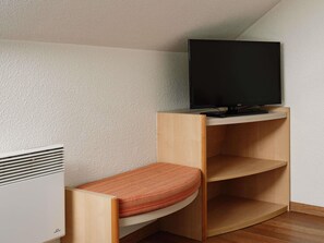 A corner room with a TV on a shelf, a bed with a patterned mattress, and a white wall.