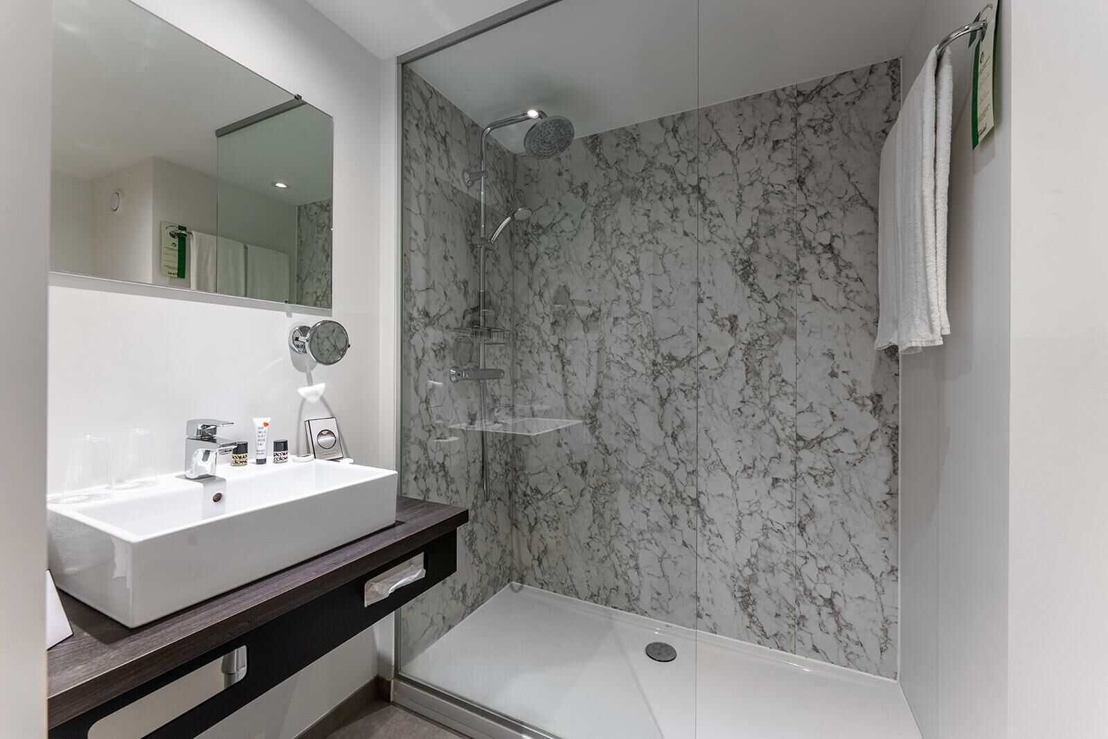 A modern bathroom with a marble shower wall, a white sink, and a large mirror.