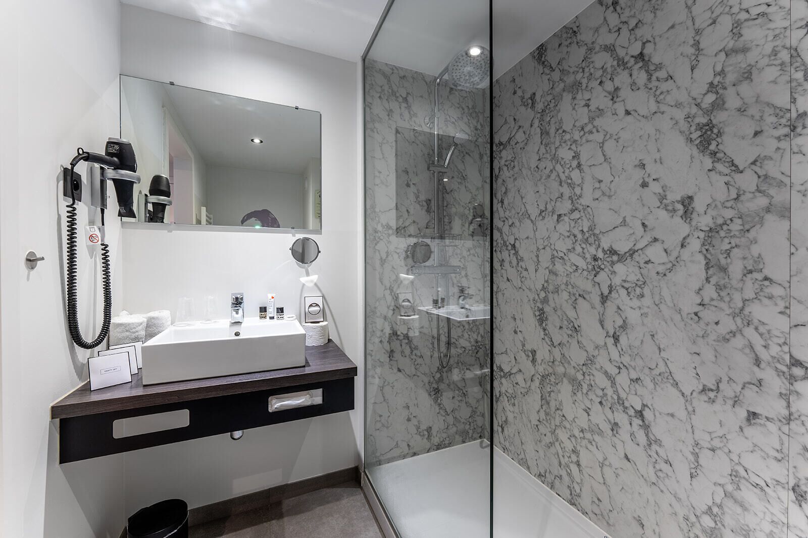 A modern bathroom with a marble shower wall, a large mirror, and a vanity with a sink and toiletries.