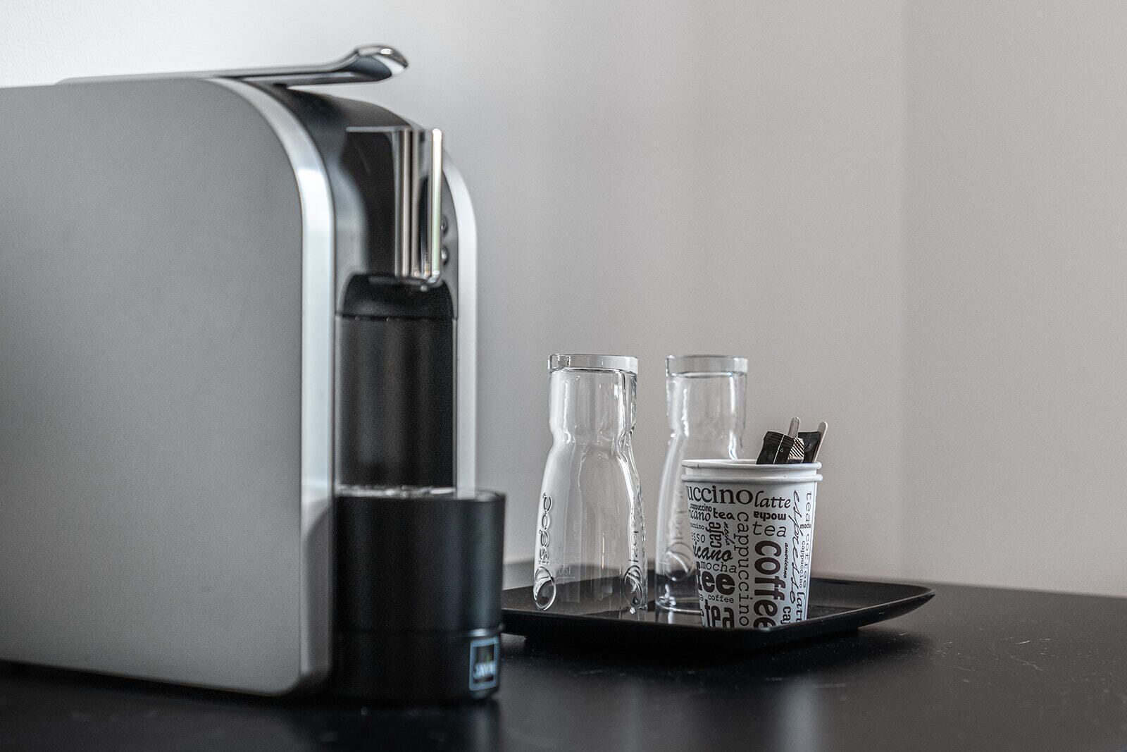 A coffee machine with a tray holding two glasses and a canister with coffee-related words.