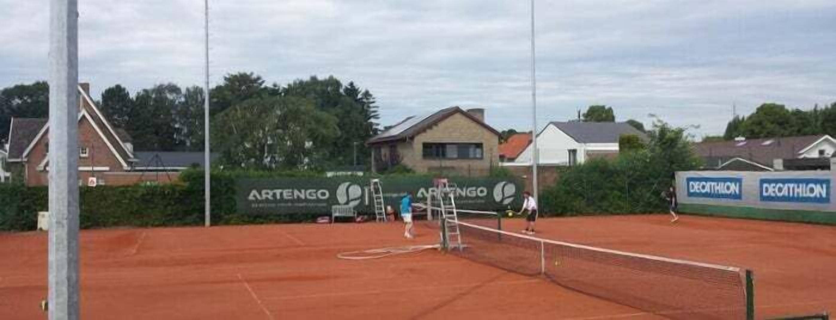 Un terrain de tennis avec un filet, entouré de maisons résidentielles et orné d’une banderole sur laquelle on peut lire le mot « ARTENGO ».
